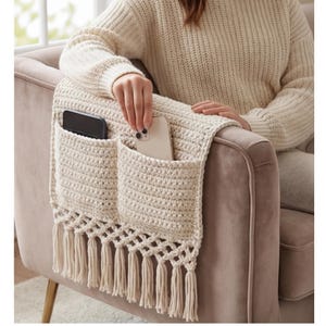 Può includere: Organizer per bracciolo all'uncinetto color crema con due tasche e frange. L'organizer è drappeggiato su una poltrona di velluto, con un telefono in ogni tasca. Il motivo all'uncinetto è visibile e le frange pendono.