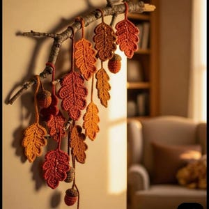 Puede incluir: Adorno de pared artesanal con temática otoñal, con hojas y bellotas de ganchillo en tonos naranja, rojo y marrón. Las hojas están suspendidas de una rama natural, creando una decoración rústica y de temporada.