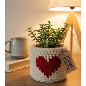 May include: A handmade crochet plant pot with a red heart design. The pot is filled with a green plant. A tag that says "HANDMADE WITH LOVE" is attached to the pot. A mug and a book are in the background.