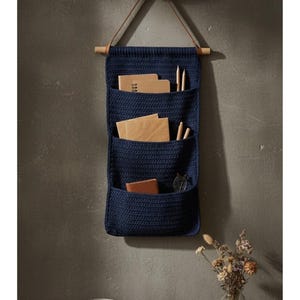 May include: A navy blue, wall-mounted organizer with three pockets. The organizer is made of a woven material and hangs from a wooden dowel with a leather strap. The pockets contain notebooks, pencils, and a small wallet.