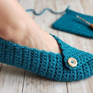 May include: Teal crochet slipper with a wooden button closure. The slipper is handmade and has a textured appearance. A matching crochet square and crochet hook are in the background.