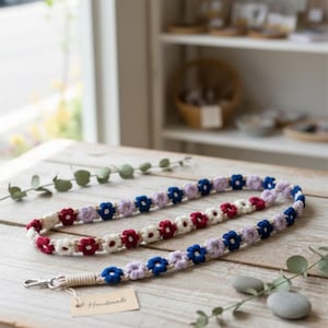 May include: A handmade lanyard with a floral design. The lanyard features crocheted flowers in red, white, blue, and lavender. The lanyard has a metal clasp and a tag that reads "Handmade".