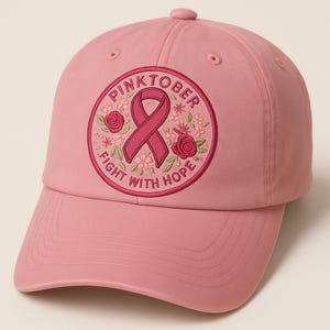 Op de afbeelding: Roze baseballpet met een geborduurde patch. De patch toont een roze lint, rozen en de woorden "PINKTOBER" en "FIGHT WITH HOPE". De pet heeft een gebogen klep en een verstelbare band.