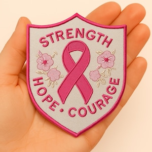Op de afbeelding: Een schildvormige patch met een roze lint, bloemen en de woorden "Strength, Hope, Courage" geborduurd in roze op een grijze achtergrond. De patch heeft een roze rand.