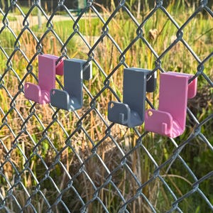 May include: Four J-shaped hooks in pink and gray are attached to a chain-link fence. The hooks are made of plastic and are designed to hang items. The background features green grass and a blurred view of a park.