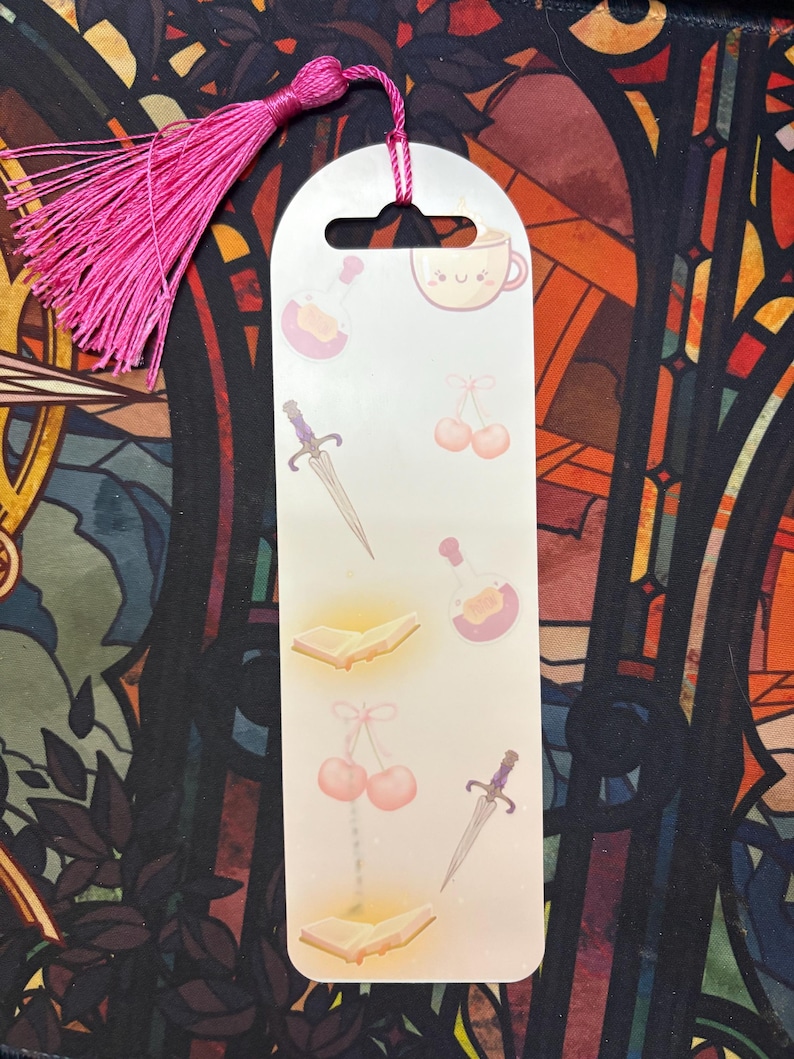 K&ouml;nnte beinhalten: Ein wei&szlig;es Lesezeichen mit einem rosa Quasten. Das Lesezeichen zeigt verschiedene Illustrationen, darunter ein Buch, einen Dolch, eine Zaubertrankflasche und Kirschen. Das Lesezeichen hat auch eine niedliche Comic-Tasse mit einem Gesicht.