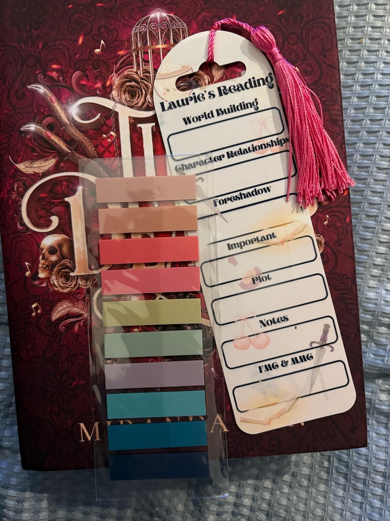 K&ouml;nnte beinhalten: Ein wei&szlig;es Lesezeichen mit einem rosa Quasten, beschriftet mit "Laurie's Reading World Building", mit Abschnitten f&uuml;r "Character Relationships", "Foreshadow", "Important Plot", "Notes" und "FMG & MMG". Das Lesezeichen ist an einer durchsichtigen Plastikfolie mit bunten Haftnotizen befestigt.