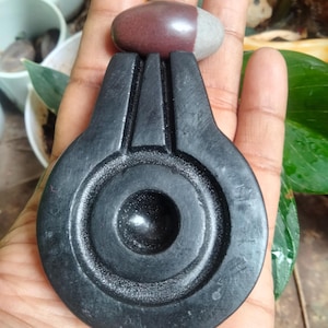 Op de afbeelding: Een zwarte stenen sculptuur met een afgeronde basis en een centraal cirkelvormig ontwerp. Een kleine, ovaalvormige steen met een donkerrode en grijze kleur rust bovenop. De sculptuur wordt in een hand gehouden.