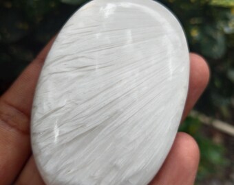 May include: A smooth, oval-shaped white stone with a fibrous, textured pattern. The stone is held in a hand, with a blurred green background. The stone's surface has a subtle sheen, and the pattern resembles fine brushstrokes.