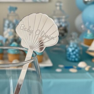 May include: A silver seashell-shaped drink stirrer with the text "Baby Oliver" is displayed in a glass. The background features a baby shower theme with blue and white balloons, cupcakes, and a small wooden sailboat.