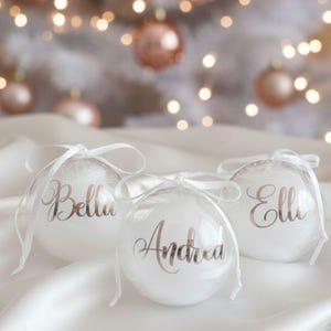 Könnte beinhalten: Drei transparente Weihnachtskugeln mit Namen in goldener Schrift: "Bella", "Andrea" und "Elle". Jede Kugel ist mit einem weißen Band gebunden. Der Hintergrund zeigt einen weißen Weihnachtsbaum mit roségoldenen Kugeln und weichen Bokeh-Lichtern.
