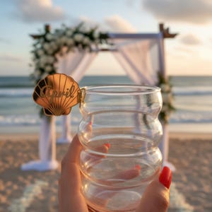 May include: A clear, bubble-shaped glass with a gold seashell charm reading "April". The glass is held up against a beach wedding backdrop with white flowers and fabric. The background shows the sea and sky.