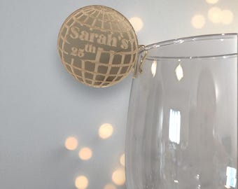 May include: A gold-colored, spherical wine charm with a globe design. The charm is attached to a clear wine glass. The text "Sarah's 25th" is etched on the charm. The background is a light gray with soft, out-of-focus lights.
