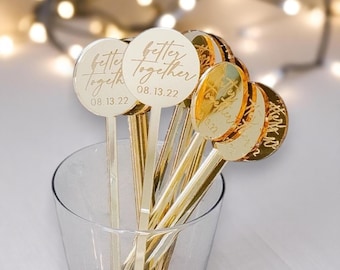 May include: Gold-colored drink stirrers with a circular top and a long stem. The text "better together" and the date "08.13.22" are engraved on the circular top. The stirrers are in a clear glass.