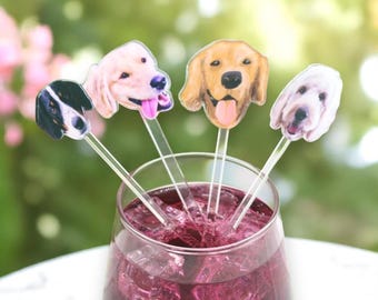 May include: A glass of red liquid with ice cubes, adorned with four dog-themed drink stirrers. Each stirrer features a different dog breed's face, including a black and white dog, a golden retriever, and a white dog.