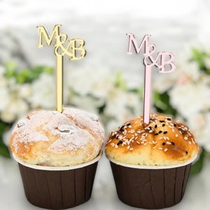 Puede incluir: Dos cupcakes con adornos decorativos. Uno tiene un adorno dorado "M&B", y el otro un adorno rosa "M&B". Los cupcakes están en moldes de papel marrón, uno espolvoreado con azúcar glas y el otro cubierto con semillas de sésamo.
