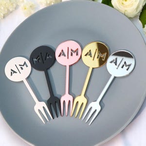May include: Five decorative cocktail forks in white, black, pink, gold, and silver, each with a circular top featuring the letters "A/M". The forks are arranged on a gray plate, ideal for a wedding or party.