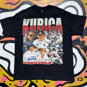 以下が含まれることがあります： ロバート・クビサのグラフィックプリントが施された黒の半袖Tシャツ。デザインには、赤と白の背景に大きな白い文字でKUBICAと書かれ、レーシングギアを身に着けたドライバーの複数の写真と、下部にGIGA KUBICAの文字が含まれています。
