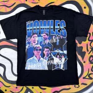 May include: A black short-sleeve t-shirt featuring a graphic print with the text VOWLES in large blue letters. The design includes multiple photographic portraits of a man in various settings, including a star-shaped inset with group photos.