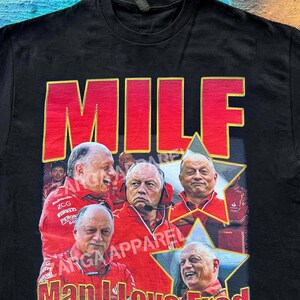 May include: A black short-sleeve t-shirt featuring a graphic print with the text MILF in large red letters at the top and Man I Love Fred at the bottom. The design includes multiple images of a man in a red jacket inside yellow-bordered stars.