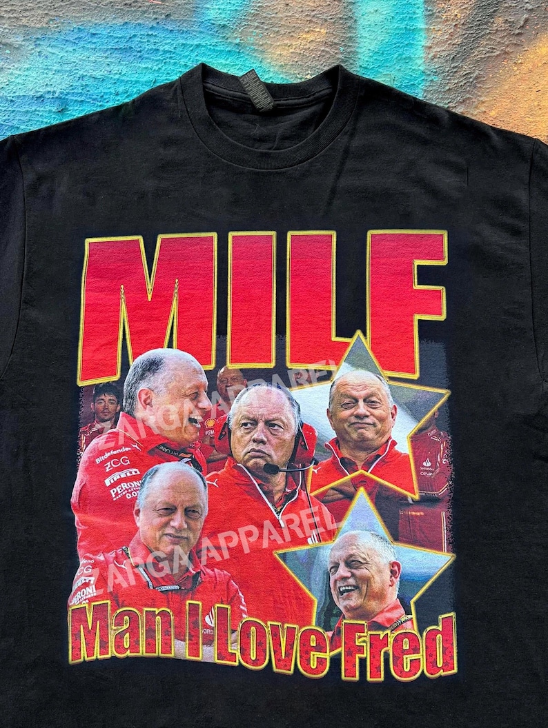 May include: A black short-sleeve t-shirt featuring a graphic print with the text MILF in large red letters at the top and Man I Love Fred at the bottom. The design includes multiple images of a man in a red jacket inside yellow-bordered stars.