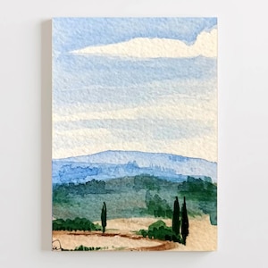 Puede incluir: Pintura de paisaje en acuarela que presenta un cielo azul con una nube blanca. La pintura representa colinas onduladas en tonos azules y verdes, con árboles y un camino en primer plano. La obra de arte está sobre papel rectangular.