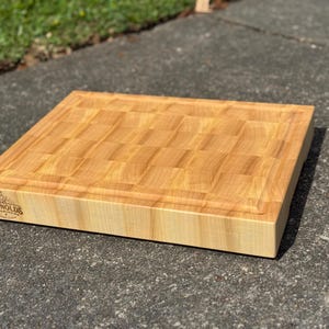 May include: A rectangular end-grain wooden cutting board with a light tan color and a grid pattern. The board features a juice groove along the top edge and a branded logo on the side that reads REYNOLDS WOODWORKING with a small icon.