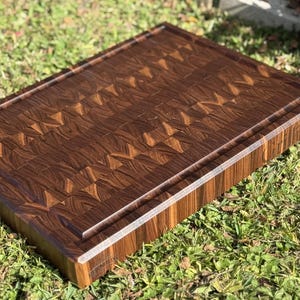 May include: A rectangular end-grain cutting board made of dark brown wood. The board features a juice groove around the perimeter and a patterned surface. The cutting board is designed for kitchen use and food preparation.
