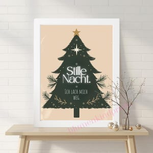 Puede incluir: Un cuadro enmarcado con un diseño de árbol de Navidad verde oscuro, con las palabras "Stille Nacht" e "Ich lach mich weg" en letras blancas y doradas. La obra de arte tiene un fondo beige y se exhibe sobre una mesa de madera.