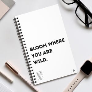 Może przedstawiać: Biały notatnik w spiralnej oprawie z napisem "BLOOM WHERE YOU ARE WILD" pogrubioną czcionką. Widoczny jest również długopis w kolorze różowego złota, czarne okulary i smartwatch. Notatnik ma również mały cytat na dole.