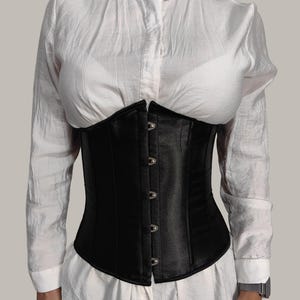 Könnte beinhalten: Ein schwarzes Satin-Korsett mit einem Busk-Verschluss und silbernen Metallösen. Das Korsett hat eine Knochenstruktur und wird über einem weißen Hemd mit Knöpfen getragen.