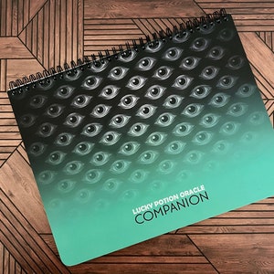 May include: A green and black spiral bound notebook with a repeating pattern of eyes. The notebook has the text "Lucky Potion Oracle Companion" printed on the front.