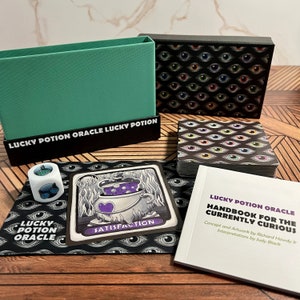 May include: A black box with a deck of cards featuring colorful eyes. The box is labeled "Lucky Potion Oracle". The cards are on a wooden surface with a green box, a white die with a blue and green eye, and a card with a purple potion in a cauldron.