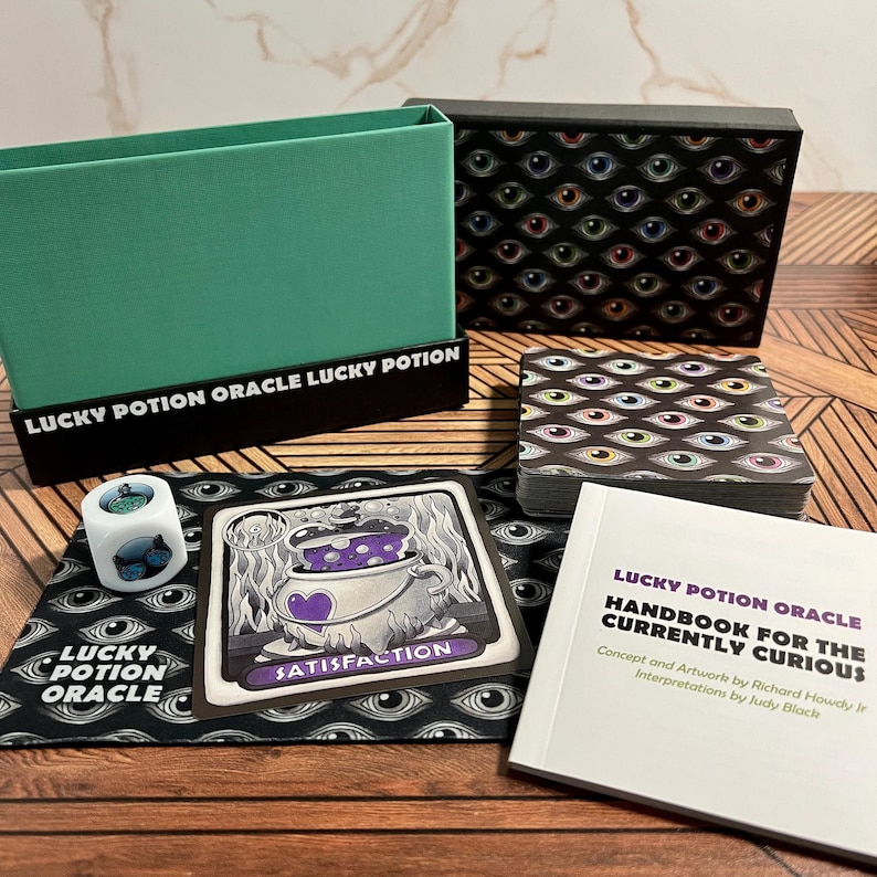 May include: A black box with a deck of cards featuring colorful eyes. The box is labeled "Lucky Potion Oracle". The cards are on a wooden surface with a green box, a white die with a blue and green eye, and a card with a purple potion in a cauldron.