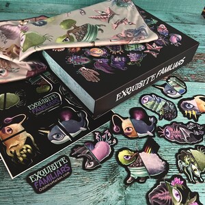 Puede incluir: Una caja negra con el texto "EXQUISITE FAMILIARS" en el lateral. La caja est&aacute; abierta y contiene un conjunto de pegatinas de colores con diversas criaturas fant&aacute;sticas. Las pegatinas est&aacute;n sobre una superficie de madera turquesa.