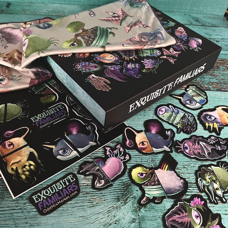 Puede incluir: Una caja negra con el texto "EXQUISITE FAMILIARS" en el lateral. La caja est&aacute; abierta y contiene un conjunto de pegatinas de colores con diversas criaturas fant&aacute;sticas. Las pegatinas est&aacute;n sobre una superficie de madera turquesa.