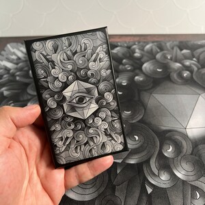May include: A black and white card with a geometric design featuring an eye inside a diamond shape. The card is surrounded by swirling clouds and a black border.