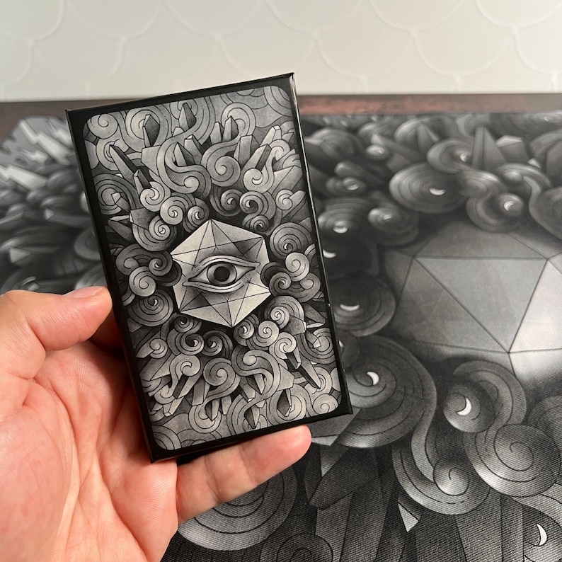 May include: A black and white card with a geometric design featuring an eye inside a diamond shape. The card is surrounded by swirling clouds and a black border.