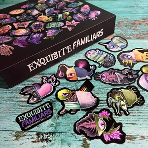 Puede incluir: Un conjunto de pegatinas con criaturas caprichosas y coloridas con detalles intrincados. Las pegatinas est&aacute;n dispuestas sobre una superficie de madera con una caja negra que dice "EXQUISITE FAMILIARS" y "OddMixMarket.com".
