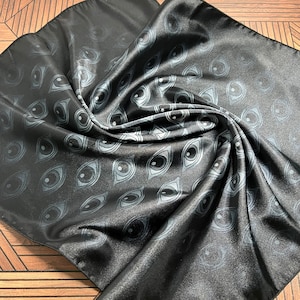 May include: A black satin scarf with a repeating pattern of white eyes. The scarf is folded and draped over a wooden surface.