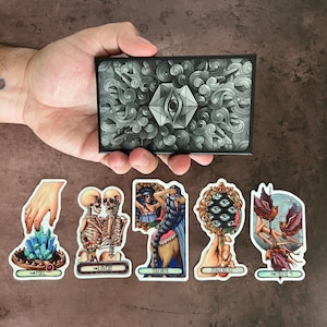 May include: Tarot card deck featuring a variety of illustrations. The cards are in a variety of colors, including black, white, red, and blue. The cards are arranged in a circle around a black and white card with a single eye in the center. The cards are labeled with the names of the suits, including The Fool, The Lovers, Strength, Judgement, and The Moon.