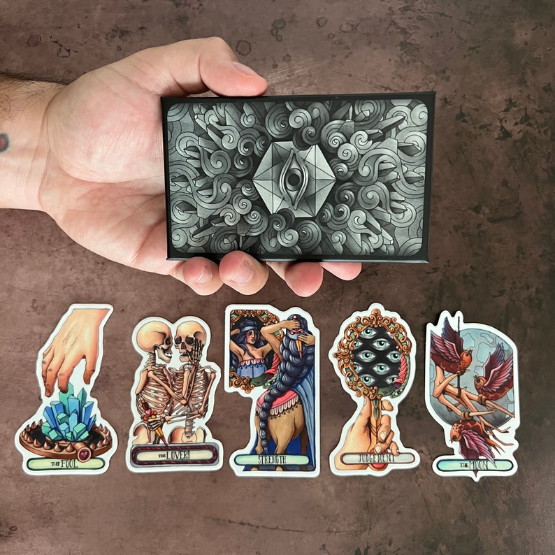 May include: Tarot card deck featuring a variety of illustrations. The cards are in a variety of colors, including black, white, red, and blue. The cards are arranged in a circle around a black and white card with a single eye in the center. The cards are labeled with the names of the suits, including The Fool, The Lovers, Strength, Judgement, and The Moon.