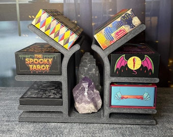 Puede incluir: Un expositor gris de varios niveles con cajas de cartas del tarot. Las cajas presentan diseños coloridos y texto, incluyendo "The Spooky Tarot". Un cristal de amatista y una piedra se encuentran en el centro.