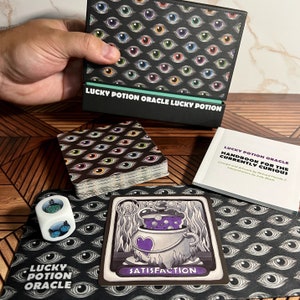 May include: A deck of oracle cards with a black background and colorful eyes. The cards are in a box with the text "Lucky Potion Oracle Lucky Potion". There is a card with a purple potion in a cauldron and the text "Satisfaction".