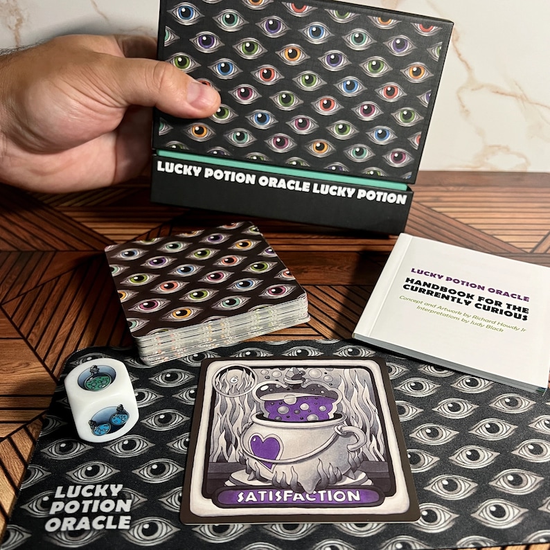 May include: A deck of oracle cards with a black background and colorful eyes. The cards are in a box with the text "Lucky Potion Oracle Lucky Potion". There is a card with a purple potion in a cauldron and the text "Satisfaction".