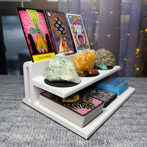 May include: A gray, tiered tarot card display stand holding several tarot cards, crystals, and decks of cards. The top tier features three tarot cards and various crystals. The bottom tier holds two decks of cards. The Six of Cups card is visible.