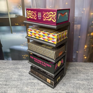 May include: A black metal display stand holding several decorative boxes. The boxes are various colors and patterns, including a box with the text "THE ART OF ADVENTURE TAROT DECK". The stand is designed to hold multiple items.