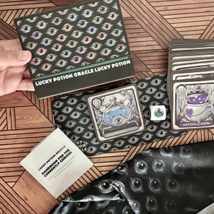 May include: A black and white oracle card deck with a black and white patterned background. The cards feature illustrations of potions and other magical items. The deck includes a handbook titled "Lucky Potion Oracle Handbook for the Currently Curious".