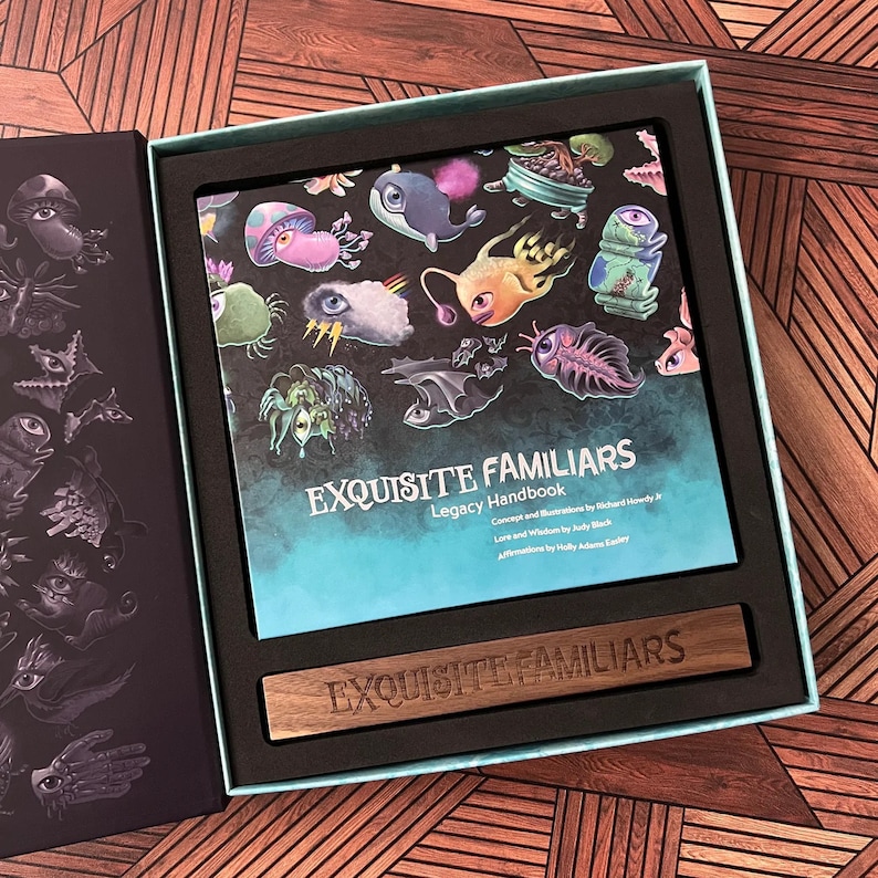 Puede incluir: Una caja azul turquesa con interior negro y una placa de madera que dice "Exquisite Familiars". La caja contiene un libro con una ilustraci&oacute;n colorida de criaturas fant&aacute;sticas en la portada. El t&iacute;tulo del libro es "Exquisite Familiars Legacy Handbook".