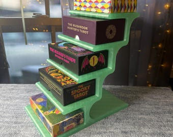 May include: A light green, tiered display stand holding five rectangular tarot card boxes. The boxes feature various colorful illustrations and titles including The Mushroom Hunter's Tarot, The Wizard's Tarot, and The Spooky Tarot, with a patterned box on top.